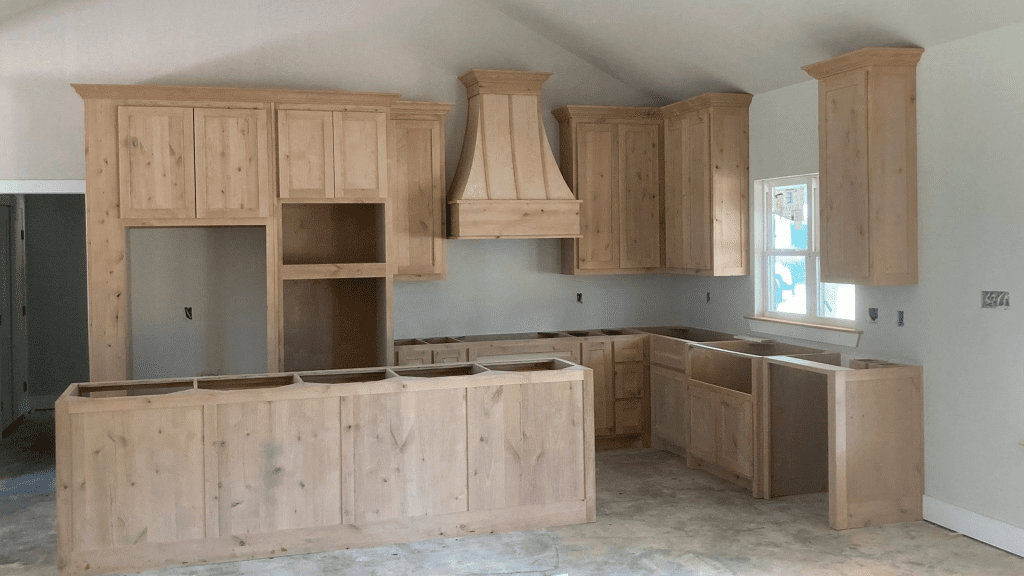 Unfinished kitchen in a custom home with light wood cabinets, empty countertop spaces, vent hood, and no appliances installed. Natural light enters through a window above the sink area.