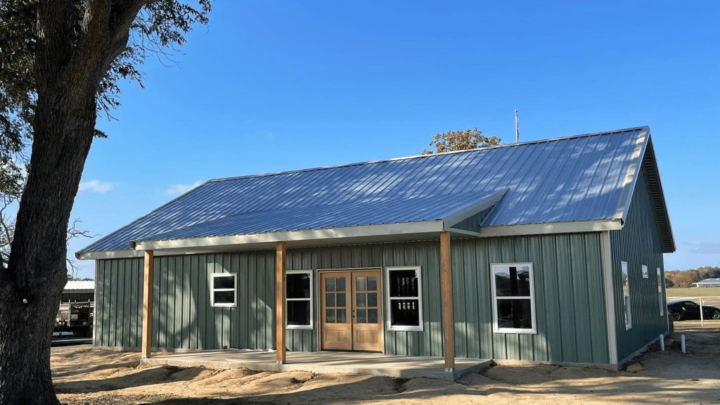 A green metal building with a sloped metal roof, wooden porch posts, and double wooden doors resembles a classic barndo, set on sandy ground under a clear blue sky.