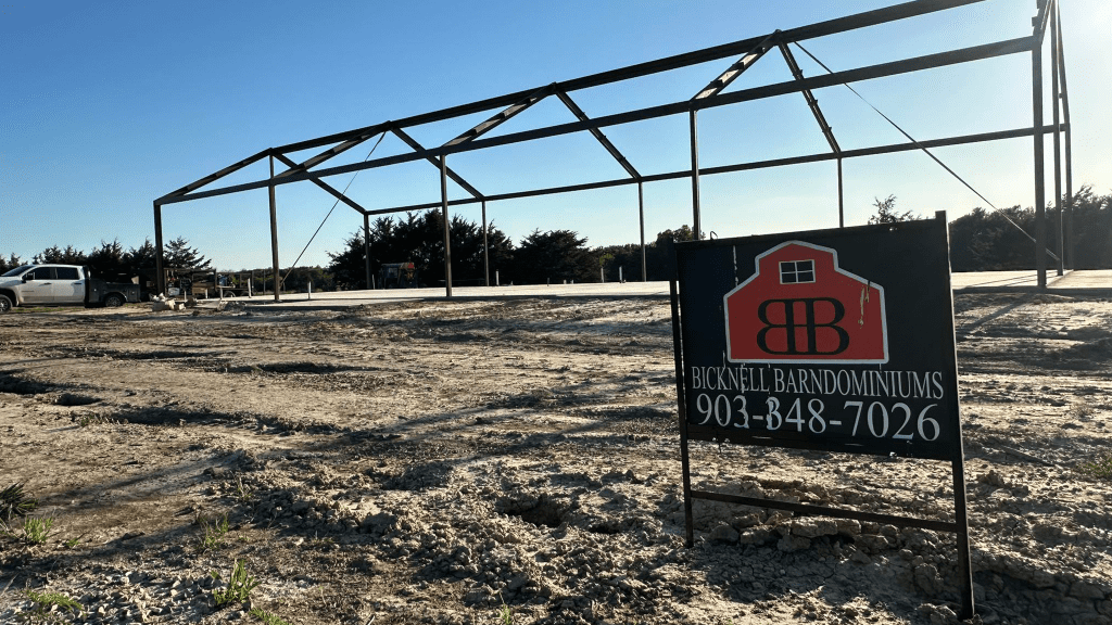 A steel frame structure is under construction on a concrete slab, with a sign in the foreground for Bicknell Barndominiums, a trusted Home Builder specializing in Barndo and Metal Shops, and a contact phone number.