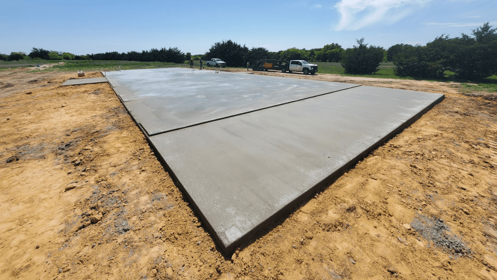 Freshly poured concrete slab foundation for a custom home on a construction site, surrounded by dirt with trucks and trees in the background under a clear sky.