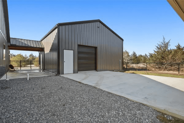 A metal garage, ideal for Metal Shops or as part of a Custom Home, features a roll-up door and side entry, connected by a covered walkway on a concrete and gravel driveway under a clear sky.