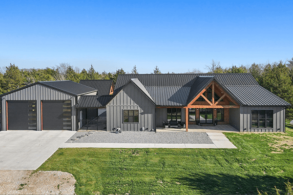 Modern gray barndo custom home with black metal roof, attached three-car garage, large windows, and covered porch, set on a grassy lot with trees in the background under a clear sky.