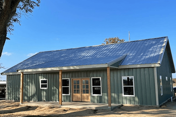 A green metal building with a sloped roof, wooden double doors, and several rectangular windows, this Barndo sits on bare ground beneath a clear blue sky.