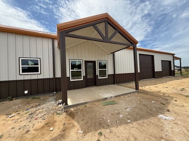 A metal barndo with a covered entryway, three windows, a door, and a large garage door sits on bare ground under a partly cloudy sky—perfect for those seeking custom home options from an experienced home builder.