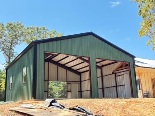 A green metal barndo with two large open garage doors stands on a dirt lot, next to a wooden structure, under a clear blue sky.