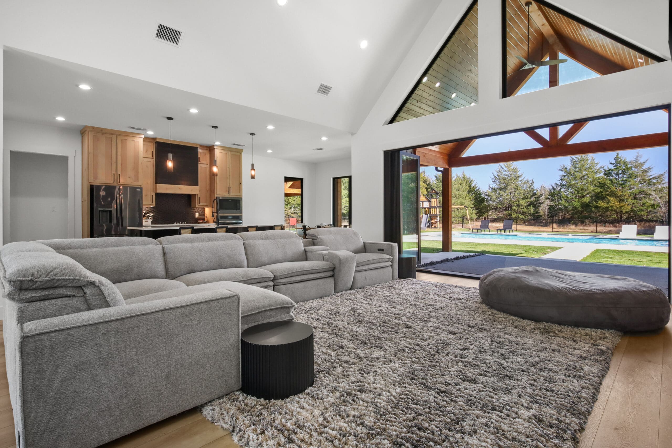 Modern living room with gray sectional sofa, textured rug, and large open glass doors leading to a patio and pool area—perfect for custom home living surrounded by trees in the background.