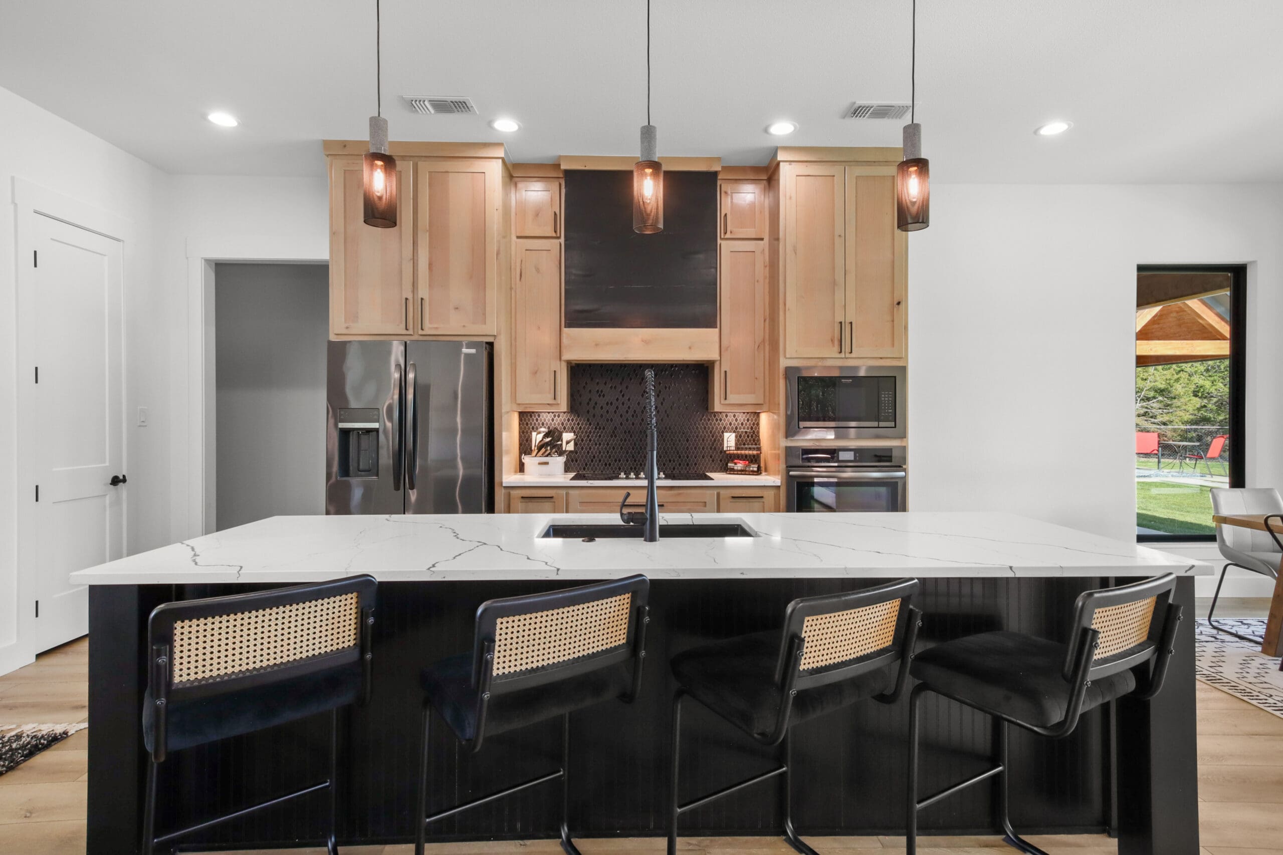Modern kitchen with a large island, four black chairs, pendant lights, stainless steel appliances, and light wood cabinets—ideal for a barndo designed by an expert home builder. Door and window reveal the outdoor view.
