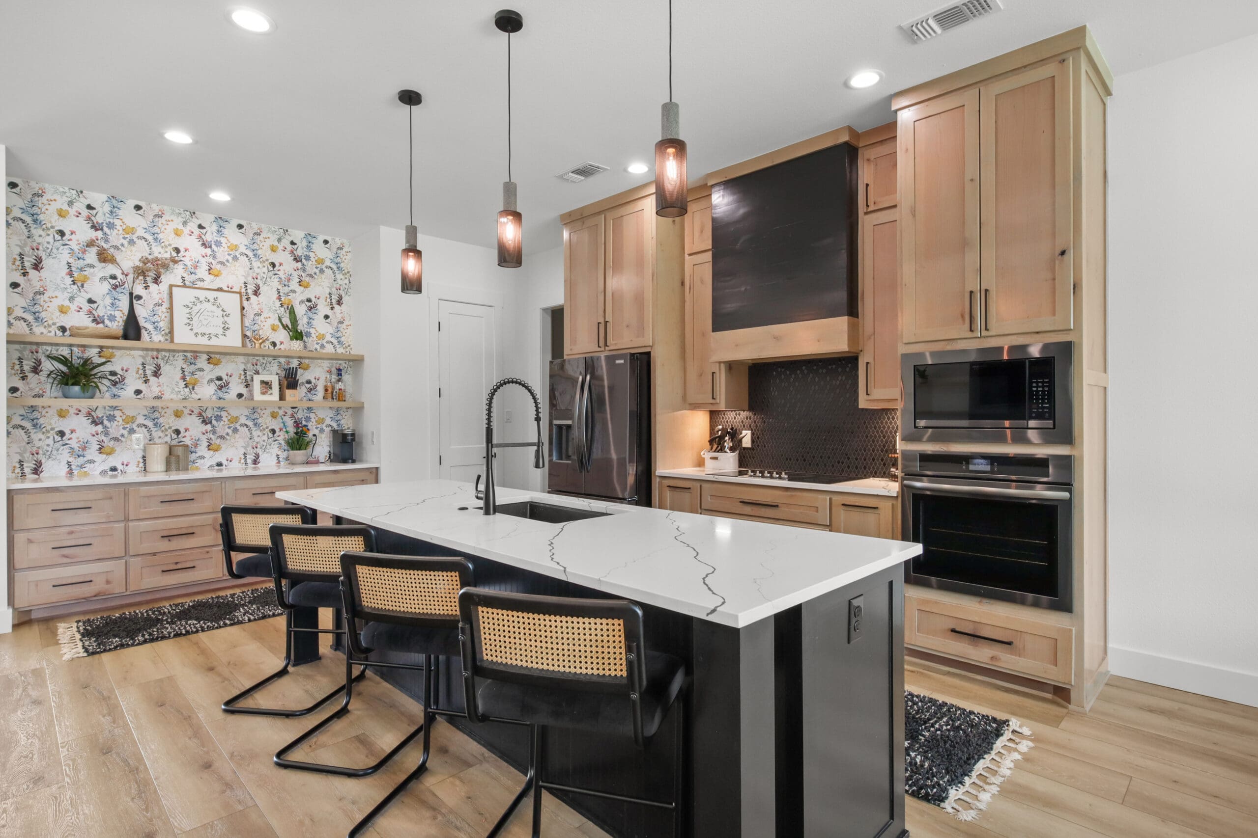 Modern kitchen by a custom home builder features light wood cabinets, black backsplash, marble island with three barstools, pendant lights, built-in appliances, and floral wallpaper on the back wall with open shelving.