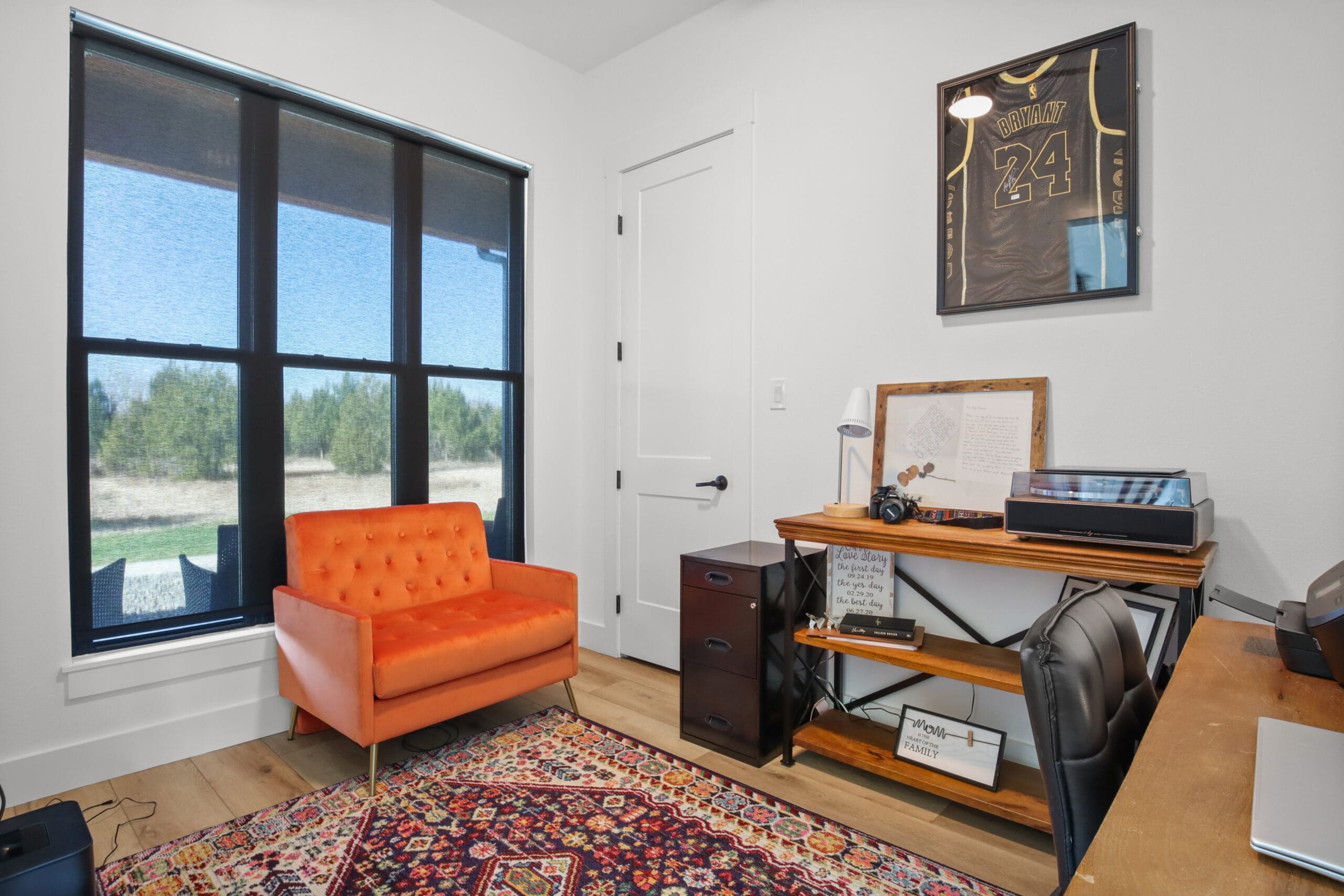 A custom home office features an orange armchair, desk, window with blackout shades, and a framed sports jersey on the wall. A colorful rug brightens the hardwood floor—perfect inspiration from a skilled home builder or Barndo design.