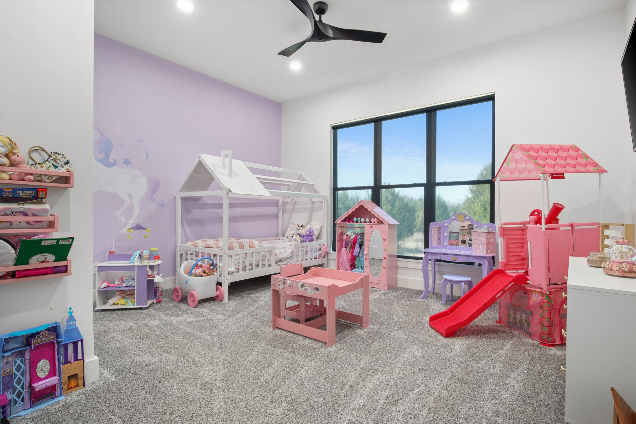 Children’s bedroom with a house-shaped bed, playhouses, toy kitchen, pink slide, toy shelves, and a large window overlooking greenery—perfect inspiration for any custom home design.