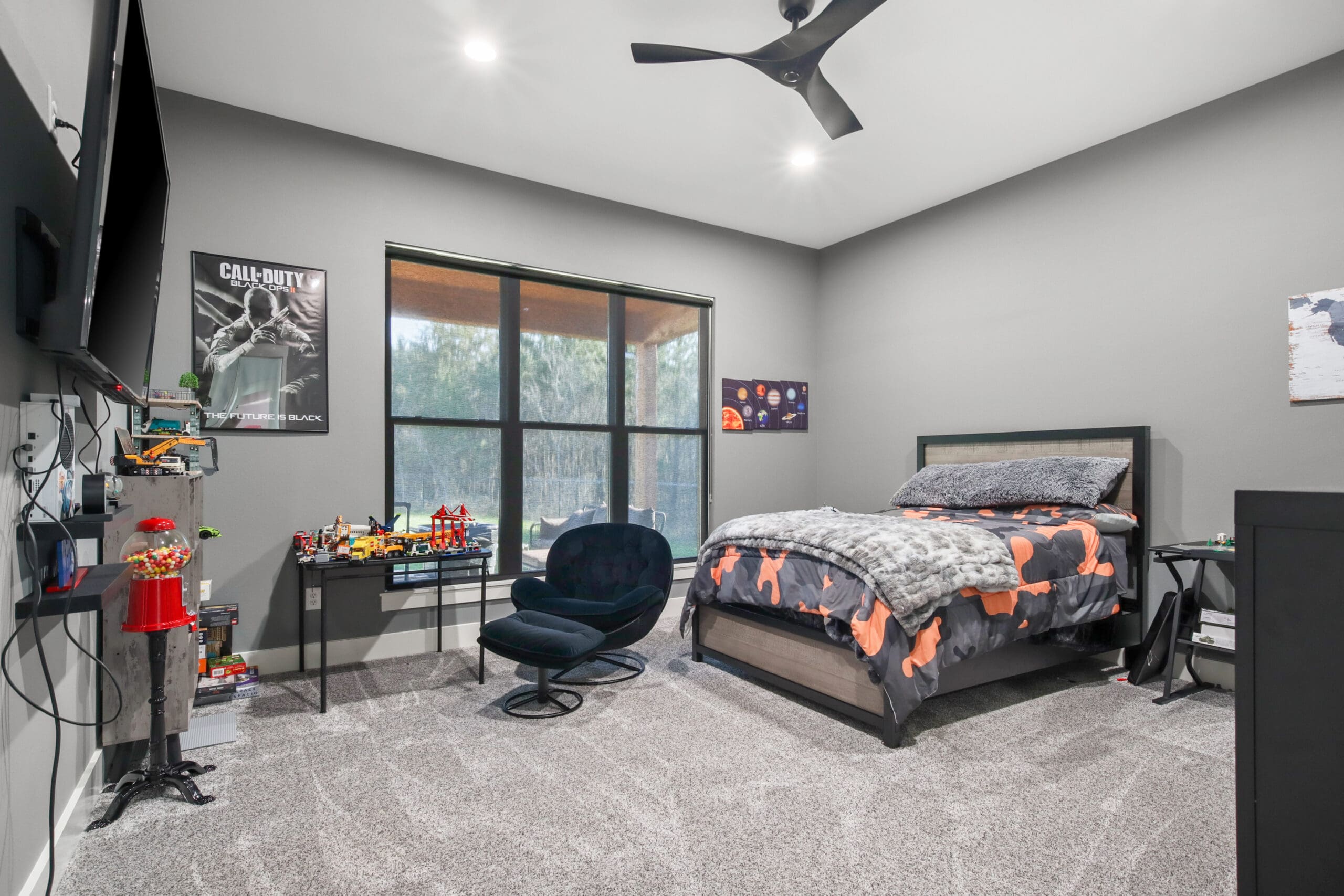 Modern bedroom in a custom home featuring a single bed, gaming chair, desk with toys, wall-mounted TV, posters, and large window with an outdoor view. Gray walls and carpet flooring add to the stylish space crafted by a skilled home builder.