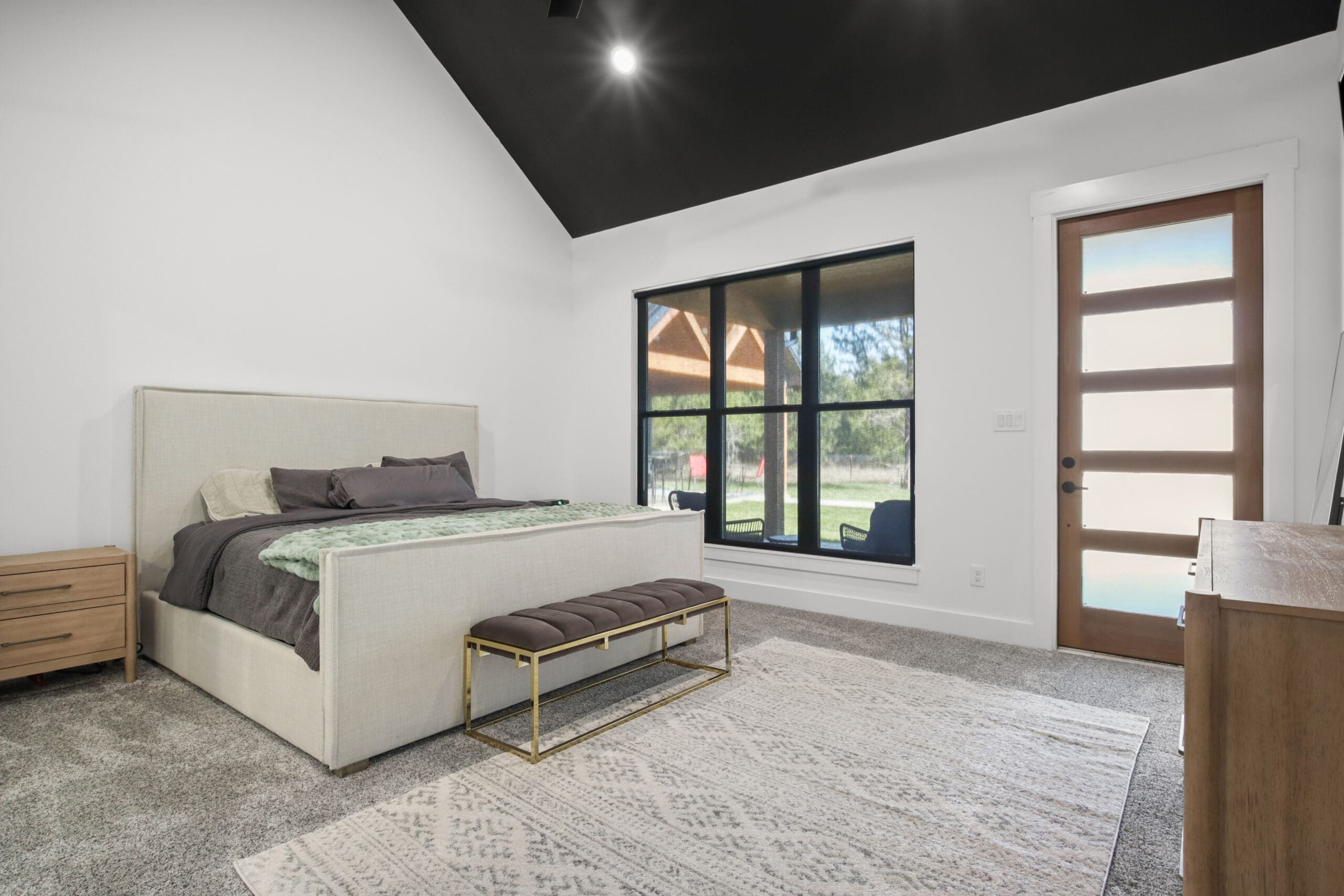 A modern Barndo bedroom features a beige bed, bench, wooden nightstand, dresser, large window, glass door, and a light-patterned rug on gray carpet. The slanted black ceiling adds character to this custom home retreat.
