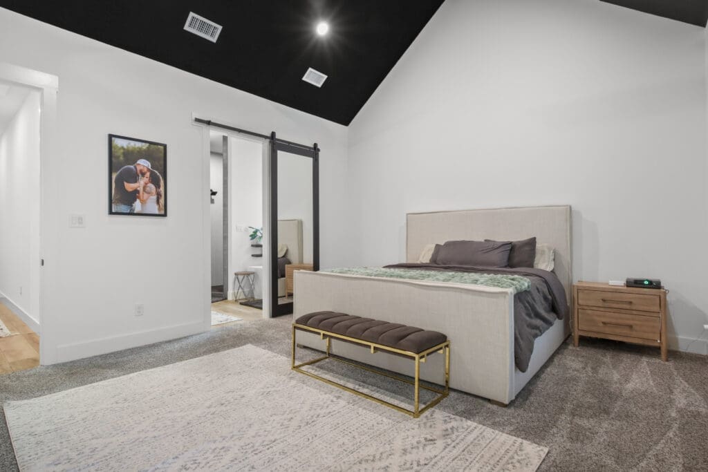Modern bedroom in a custom home features a beige upholstered bed, wooden nightstands, bench at the foot, large rug, and a sliding barn door—perfect for your barndo or metal shops-inspired retreat.