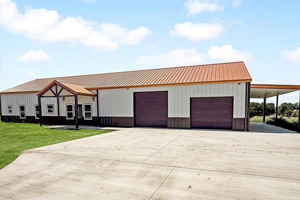 A large metal building with brown trim, two big garage doors, a covered entryway, and a concrete driveway—this custom home by an experienced home builder sits on a grassy lot under a partly cloudy sky.