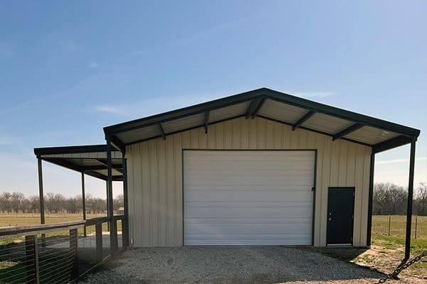 A barndo with a large white garage door, black entry door, and covered side porch stands on a gravel driveway in a rural area under a clear sky—a perfect example of what your custom home builder can create.
