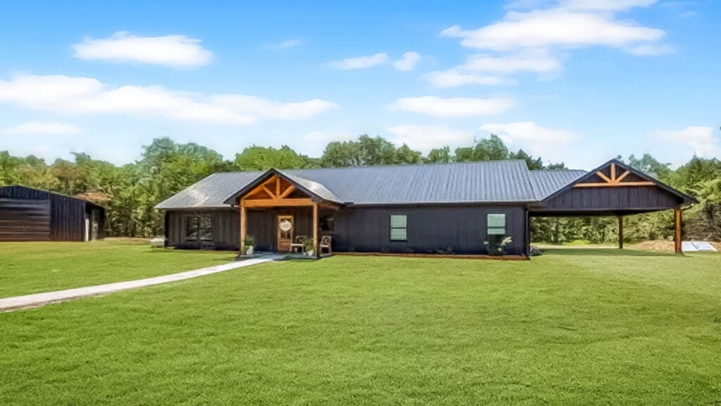 Single-story house with black siding, metal roof, a covered entryway, and attached carport, set on a large grassy lawn with trees in the background under a blue sky.