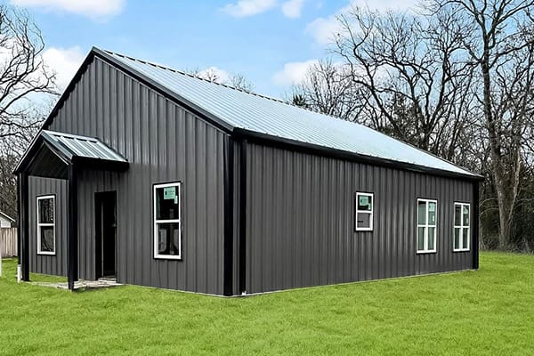 A single-story, dark gray metal barndo with a sloped roof, multiple windows, and a small covered entry, situated on a grassy lawn with leafless trees in the background.