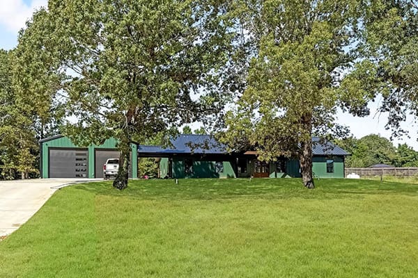 A green single-story custom home with a dark roof sits behind large trees and a mowed lawn; on the left, a detached garage with an open bay and a white vehicle completes this inviting home.