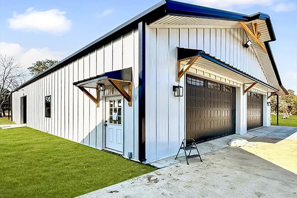 A white metal barndo with large brown garage doors, a small entry door, and a step ladder placed on a concrete driveway beside green grass—a unique design from your trusted custom home builder.