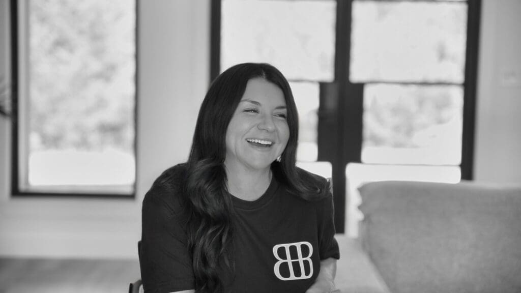 A woman with long dark hair smiles while seated indoors, wearing a dark t-shirt with a white double-B logo—perfect attire for a custom home or barndo enthusiast. Large windows and a sofa are visible in the background.