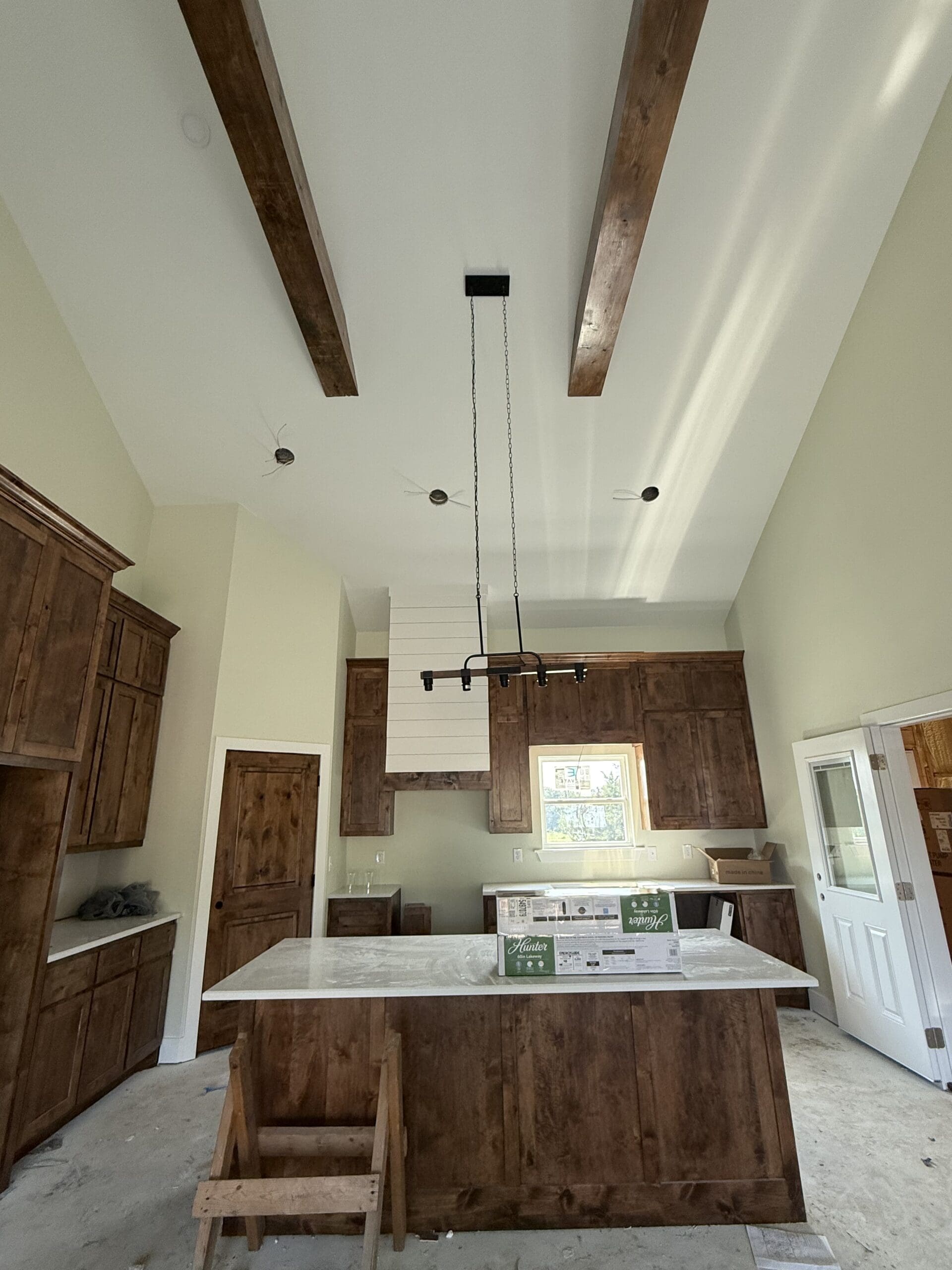 A kitchen under construction in a custom home features wooden cabinets, a marble island, exposed ceiling beams, a modern light fixture, and boxes on the countertop.