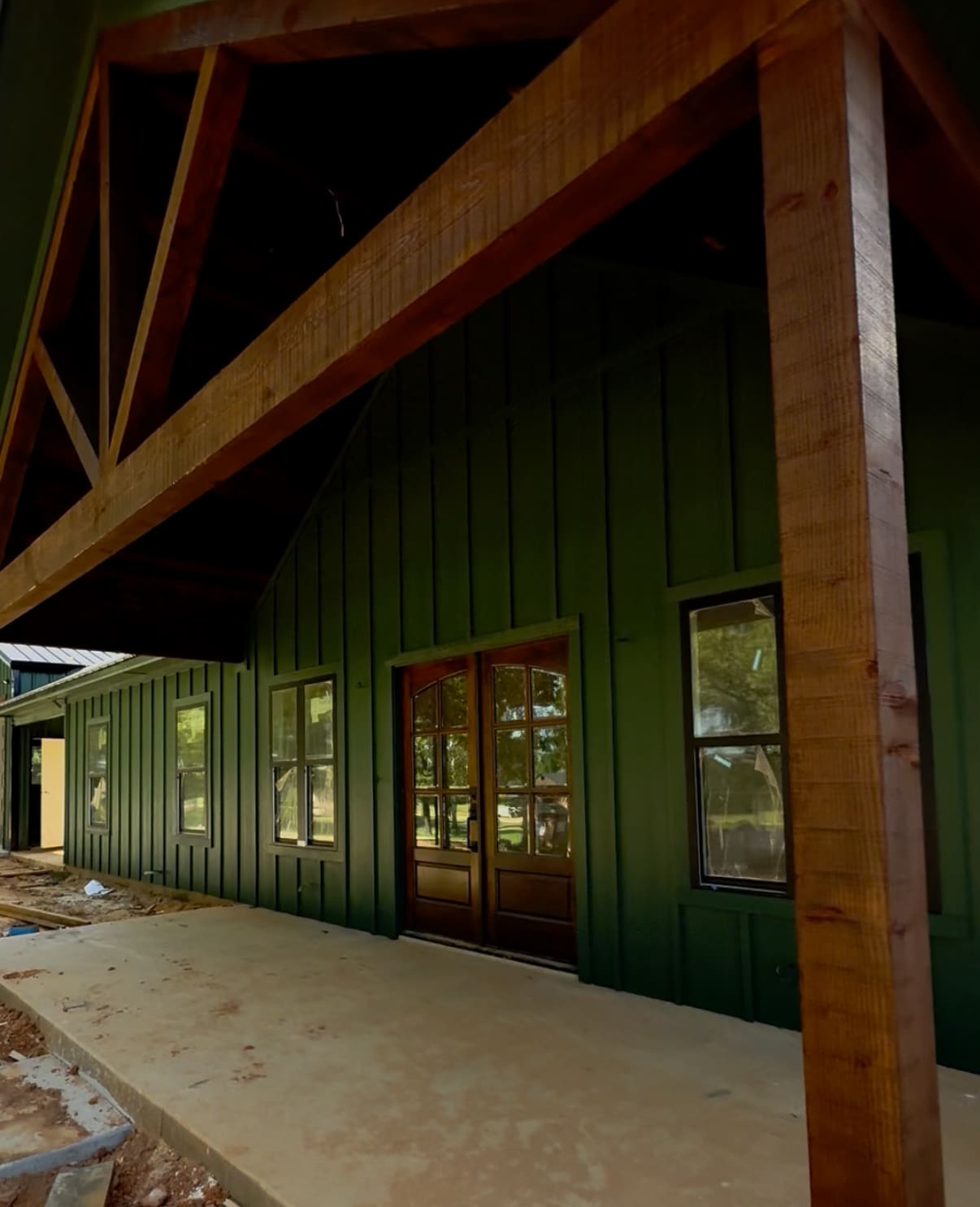 A covered porch with wood beams and green board-and-batten siding gives this custom home a welcoming touch, featuring double front doors and several windows—blending classic style with modern barndo inspiration.