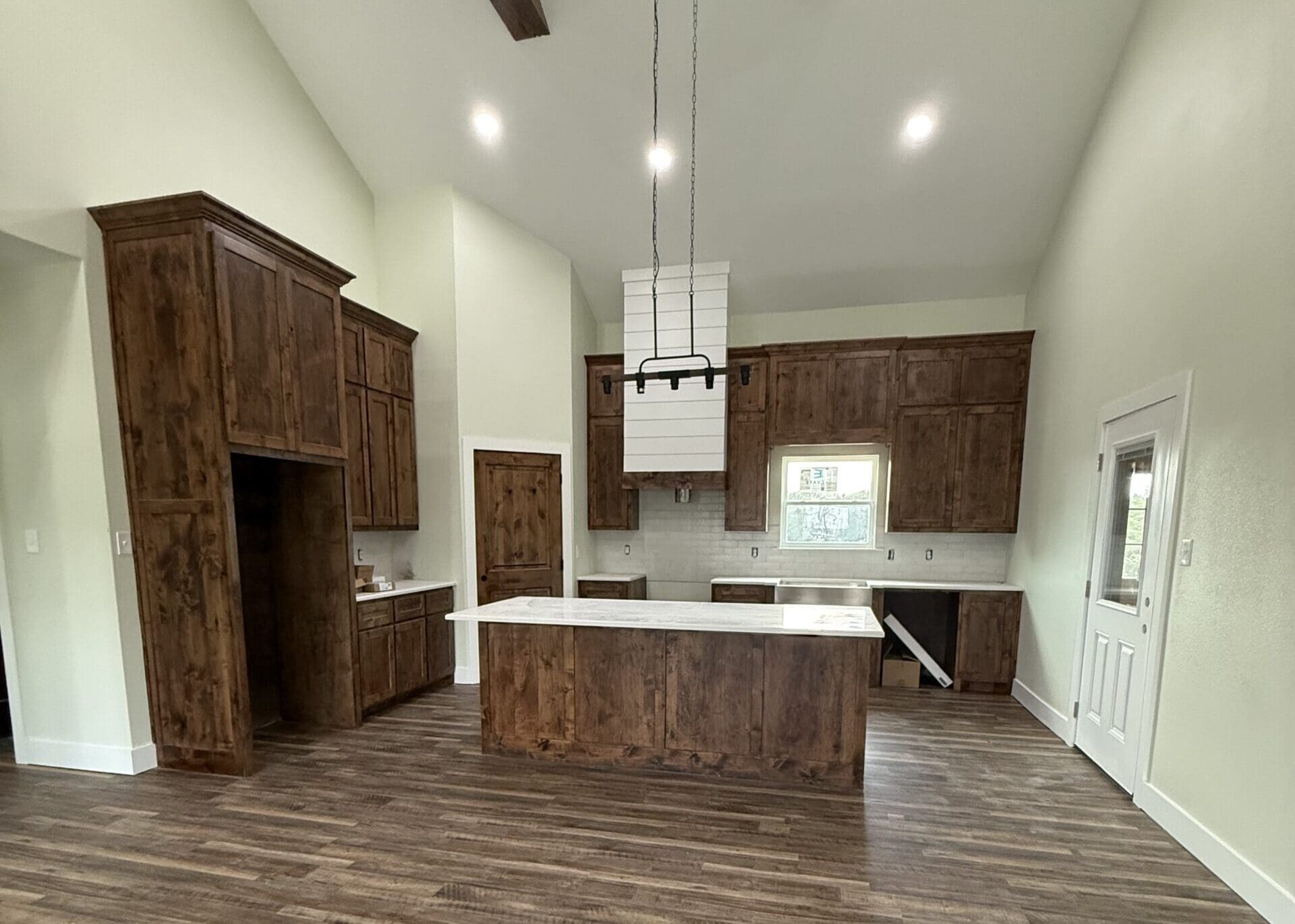 Modern kitchen in a custom home featuring dark wood cabinets, white countertops, an island, pendant lighting, white tile backsplash, and wooden floors. One cabinet door under the sink is open.
