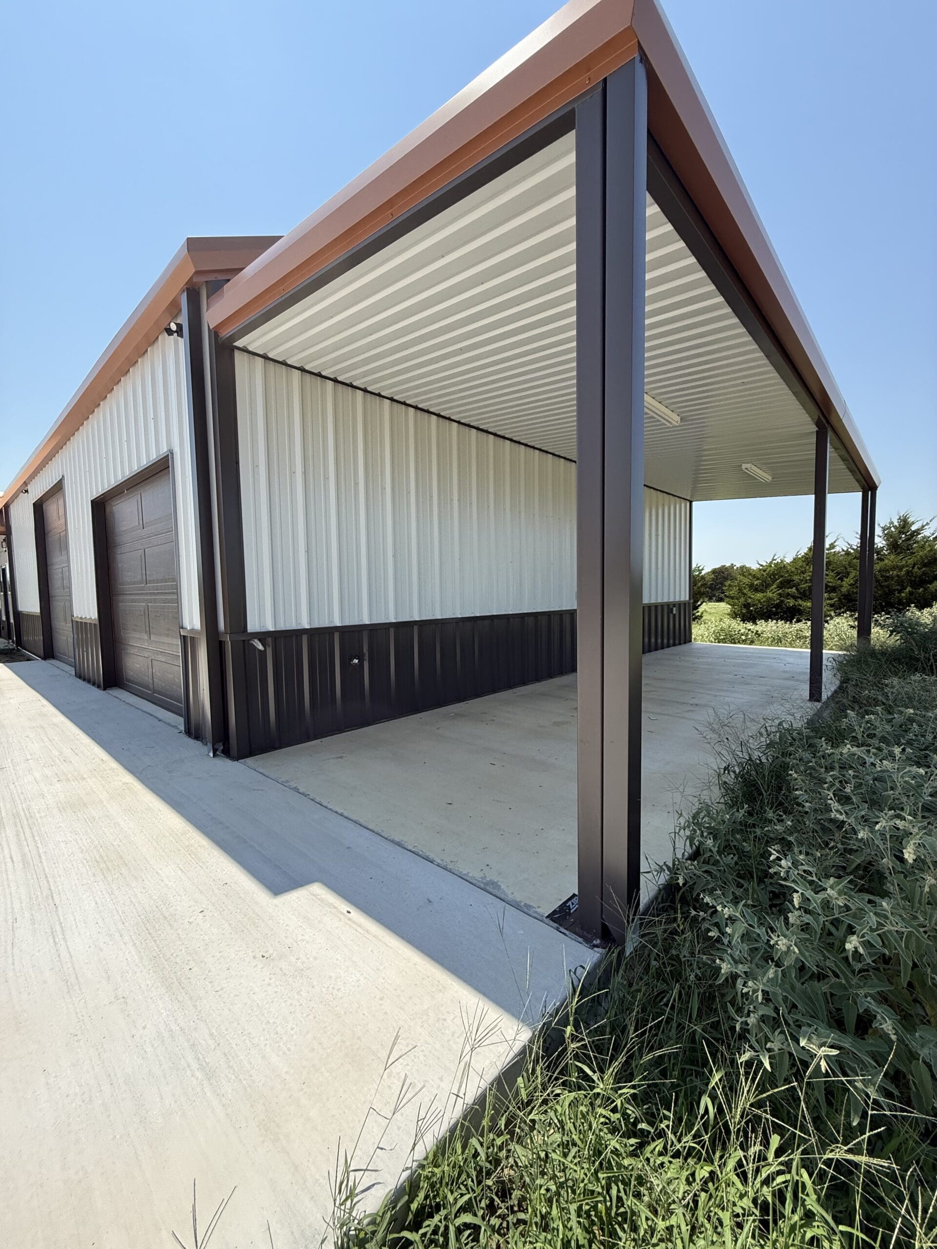 A metal carport structure attached to a large building with a garage door, featuring a concrete floor and open sides, sits beside a lush grassy area—ideal for those considering Barndo living or adding Metal Shops by your trusted Home Builder.