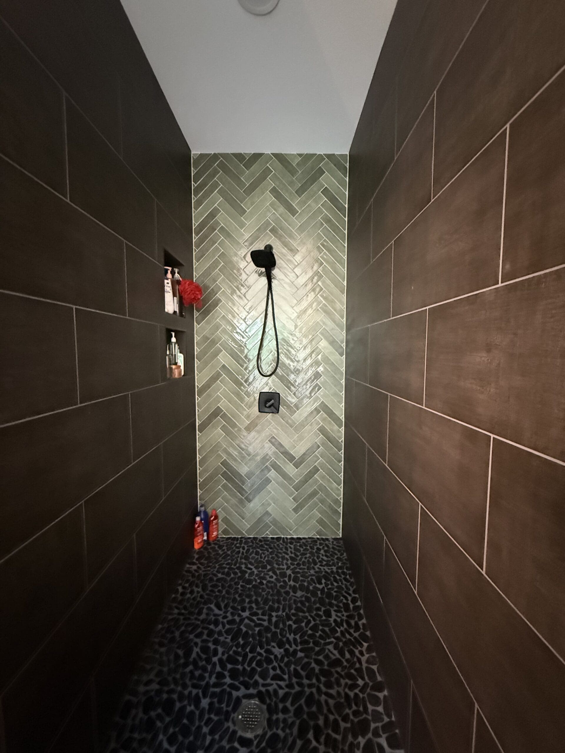 Modern shower with dark brown rectangular wall tiles, a green herringbone accent wall, pebble stone floor, and a black shower fixture—perfect for a Barndo or Custom Home—plus built-in shelf with toiletries for added convenience.