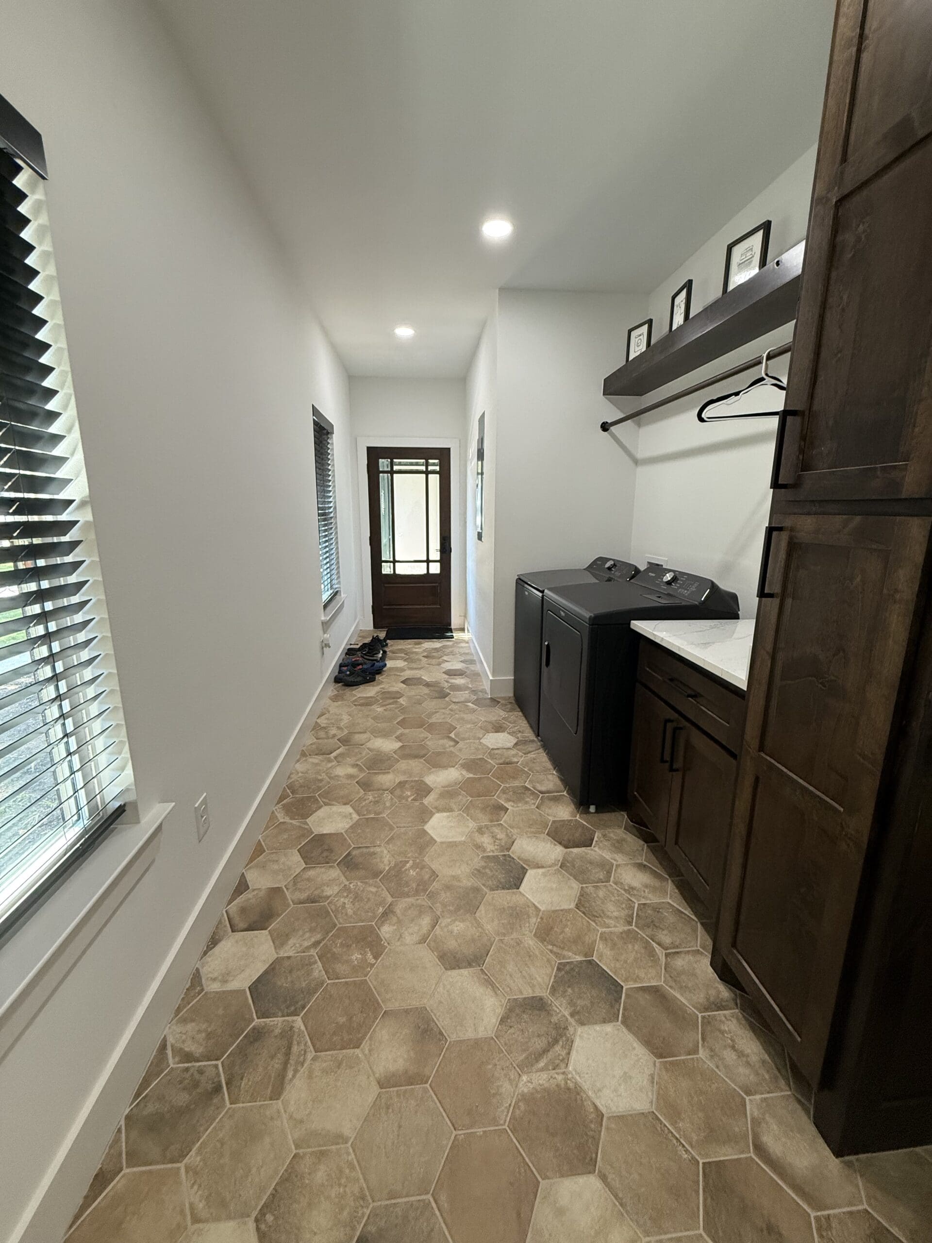 A narrow laundry room in a custom home with hexagonal tile flooring, a washer and dryer, wooden cabinets, a shelf with picture frames, two windows, and a door at the end.