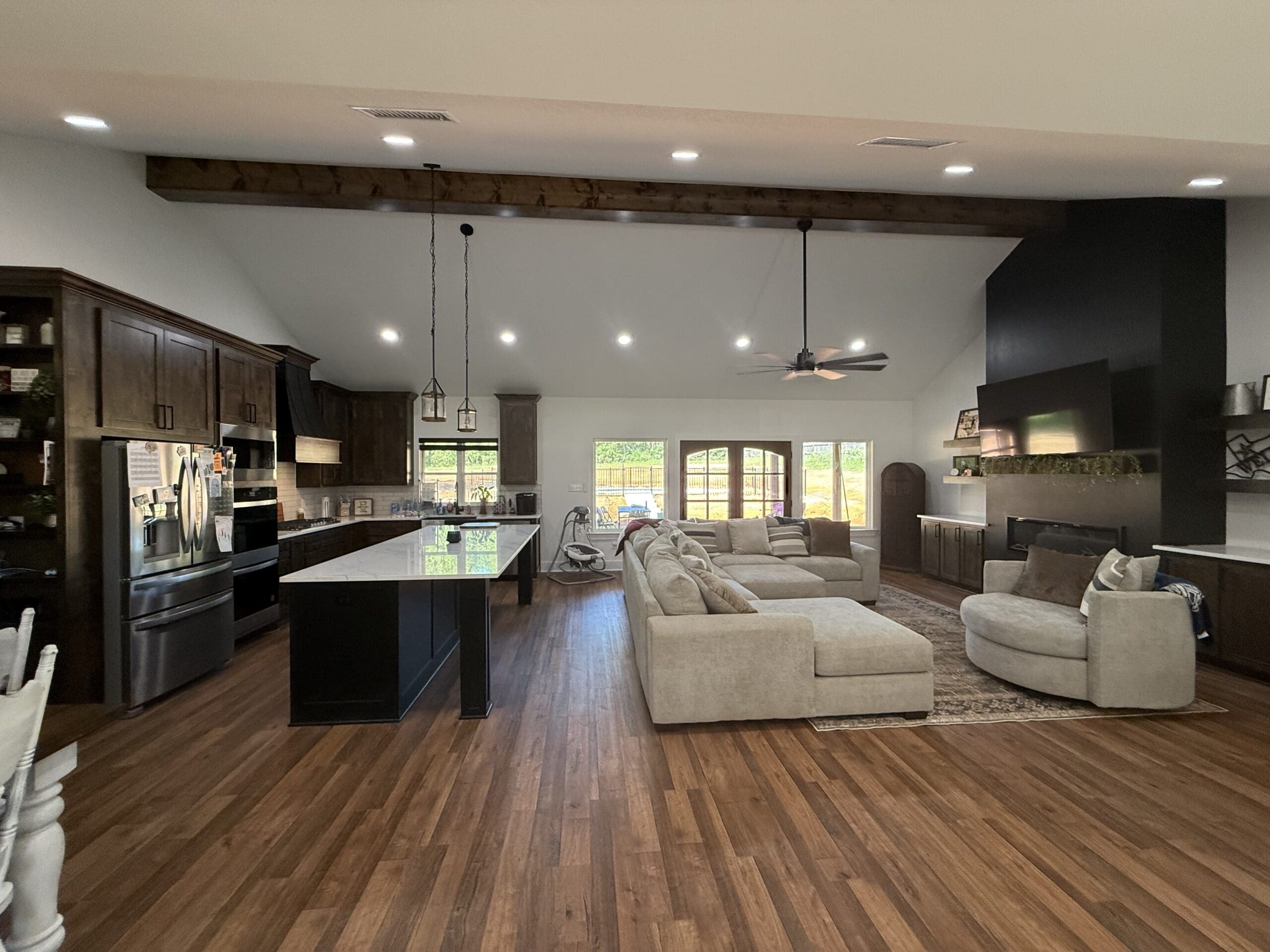 Open-concept kitchen and living room in this custom home feature dark wood floors, an island with bar stools, a beige sectional sofa, fireplace, and large windows that fill the space with natural light.