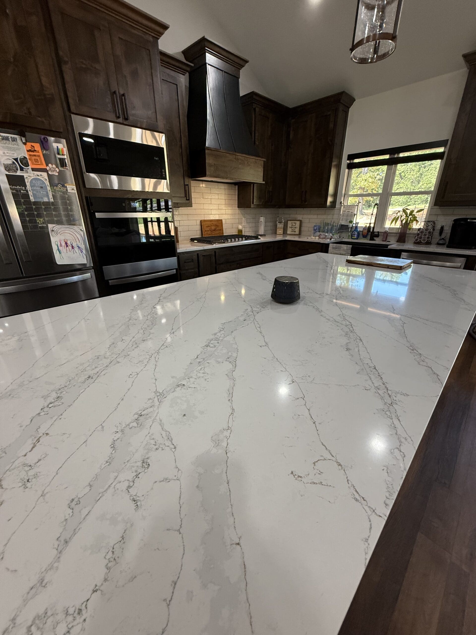 A modern kitchen in this custom home features dark wood cabinets, stainless steel appliances, white marble-patterned countertops, and a large island with a small black object at its center.