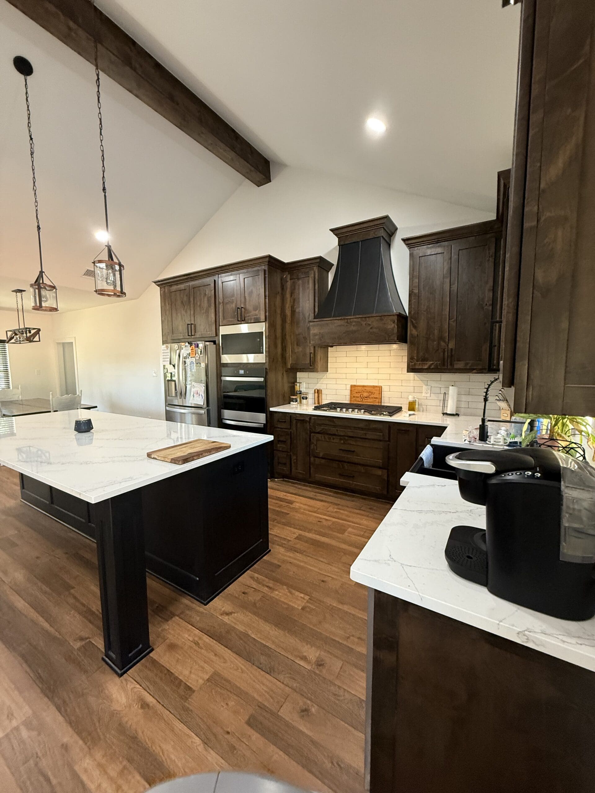 Modern kitchen in a custom home features dark wood cabinets, white marble countertops, a central island, stainless steel appliances, and pendant lighting. The wooden floor and high ceiling with exposed beam add barndo-inspired charm.
