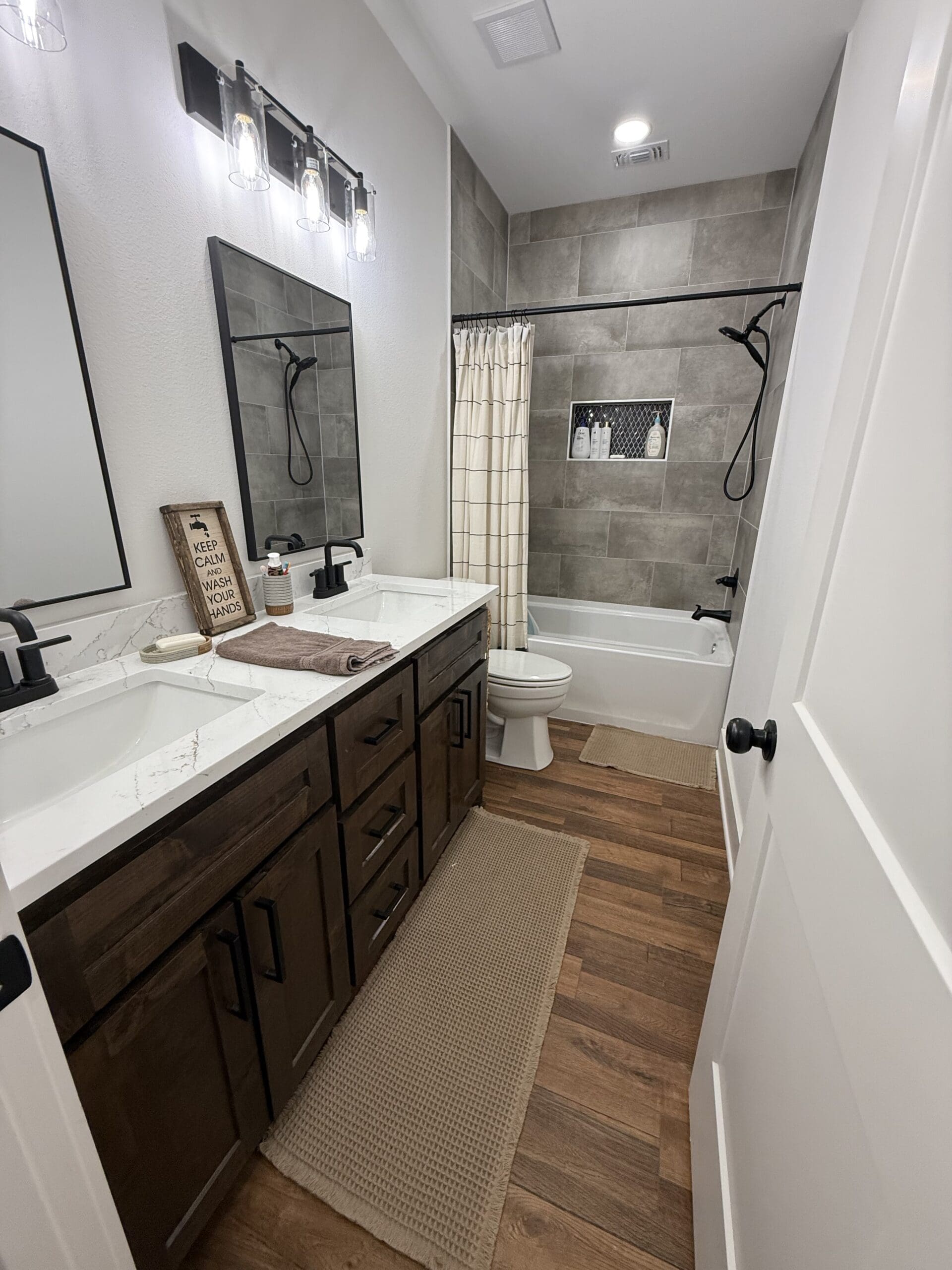 Modern bathroom in a custom home features a double-sink vanity, marble countertop, dark wood cabinets, bathtub with gray tile surround, shower curtain, and wood-look flooring—crafted by an expert home builder.