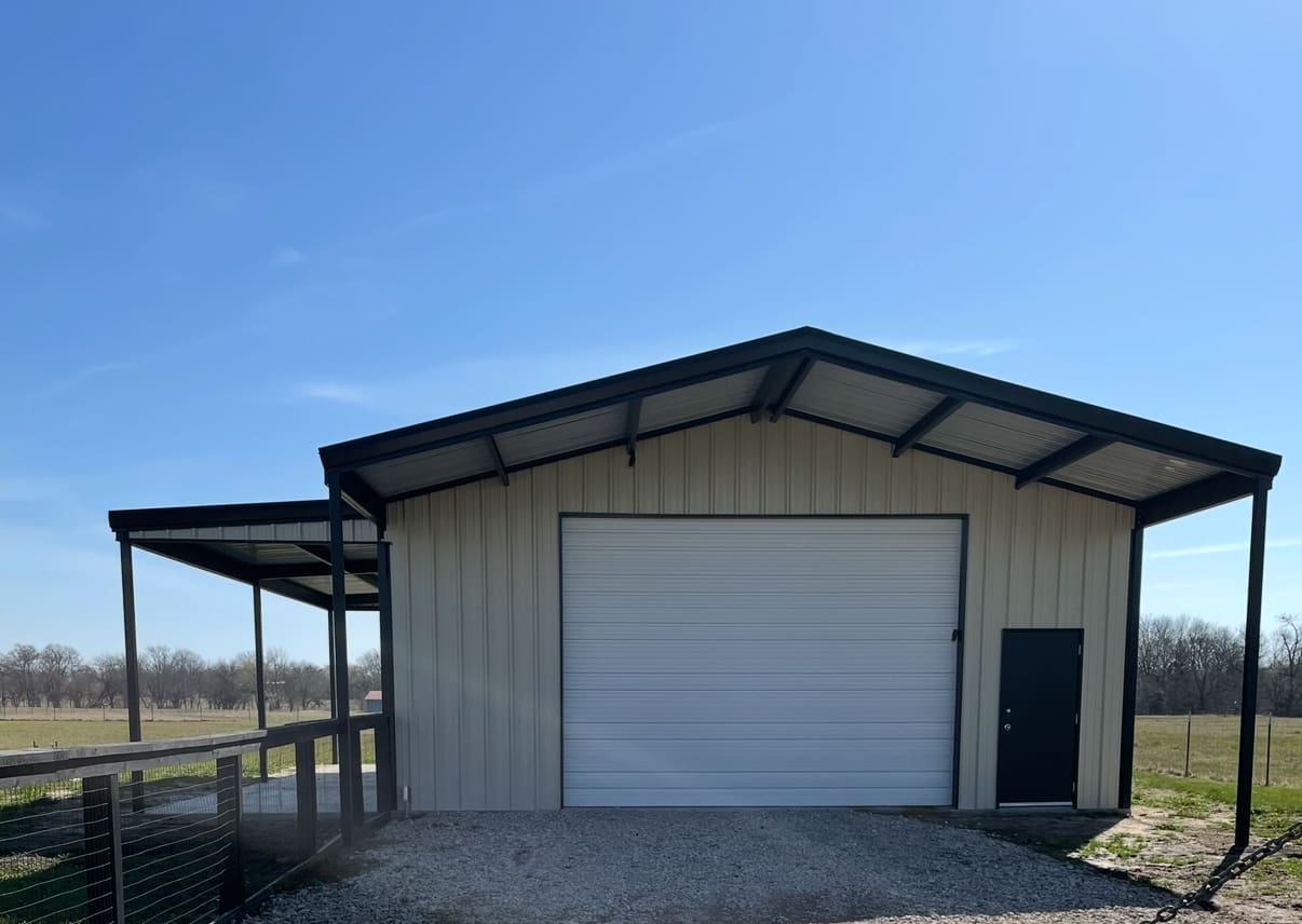 A metal building with a large white garage door, black entry door, and an attached covered area—a perfect Barndo—situated on a gravel driveway next to a fenced field under a clear sky.
