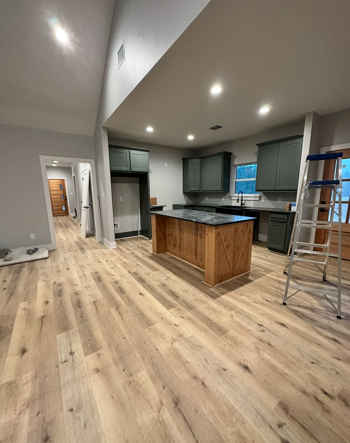 Modern kitchen with wooden floors, green cabinets, a central island with wood paneling designed by a custom home builder, and a metal ladder; hallway and closed doors visible in the background.
