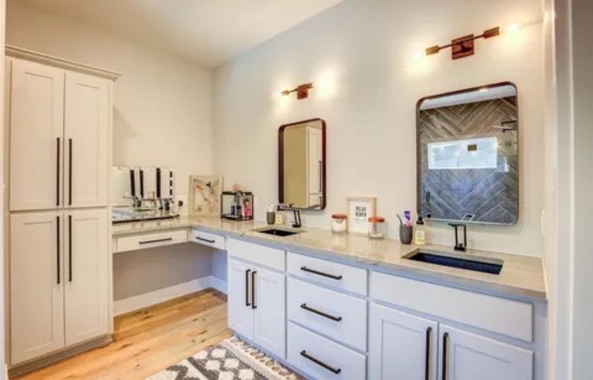 Modern bathroom by a custom home builder features double sink vanity, two mirrors, wall-mounted lights, light cabinets, wooden floor, and a chevron-tiled shower visible in the background.