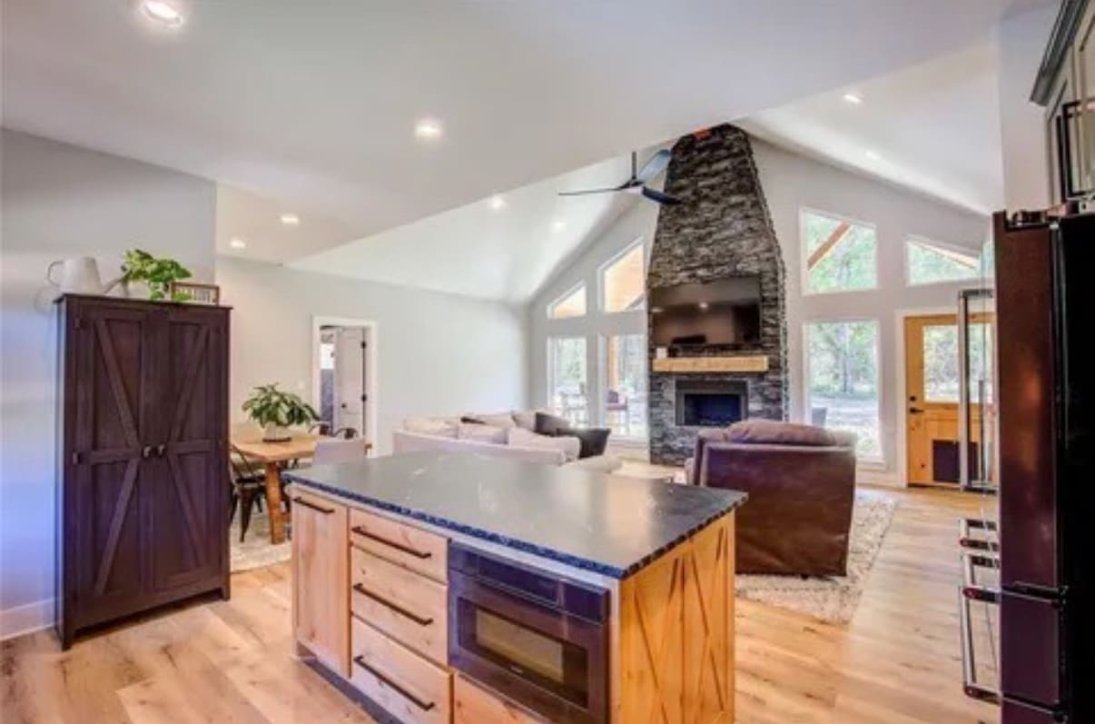Open-concept living area by a skilled home builder, featuring a kitchen island, dining table, seating area with a stone fireplace, and large windows—ideal for barndo or metal shop-inspired designs.