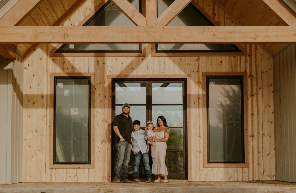 A family of four stands in front of a modern barndo-style wooden house with large windows and exposed beams.