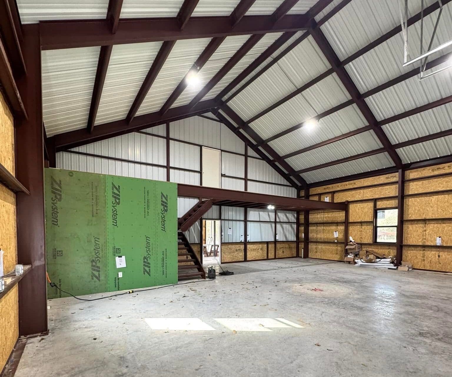 Interior of a large unfinished metal building with exposed beams, a mezzanine level, plywood walls, and a green wall panel leaning on the floor—perfect for Barndo enthusiasts or those seeking inspiration for their next Metal Shops project.