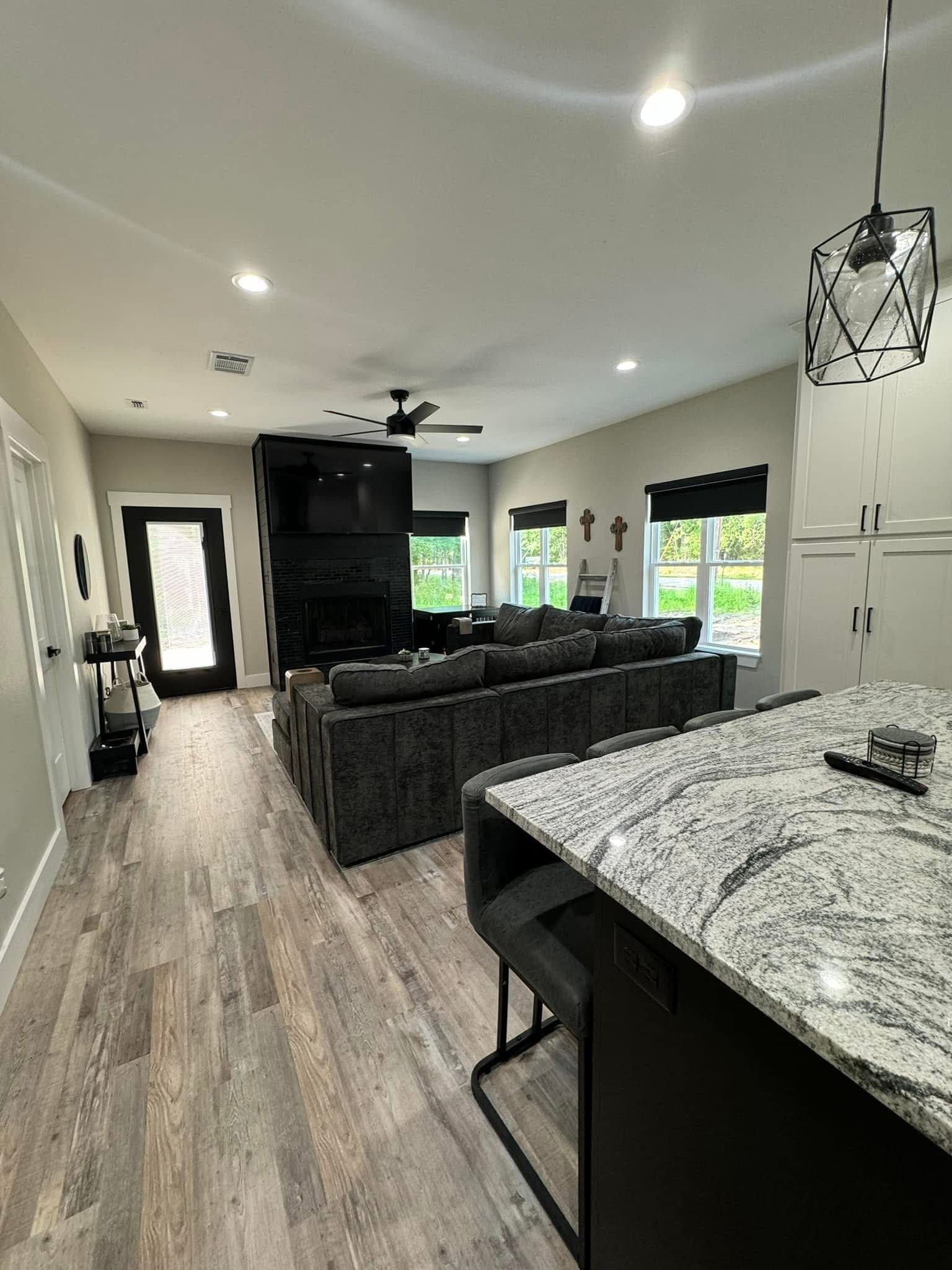 Modern living room in a custom home featuring a gray sectional sofa, wall-mounted TV, large windows, and a view into the kitchen with granite countertops and bar stools. Wood flooring runs throughout this stylish space.