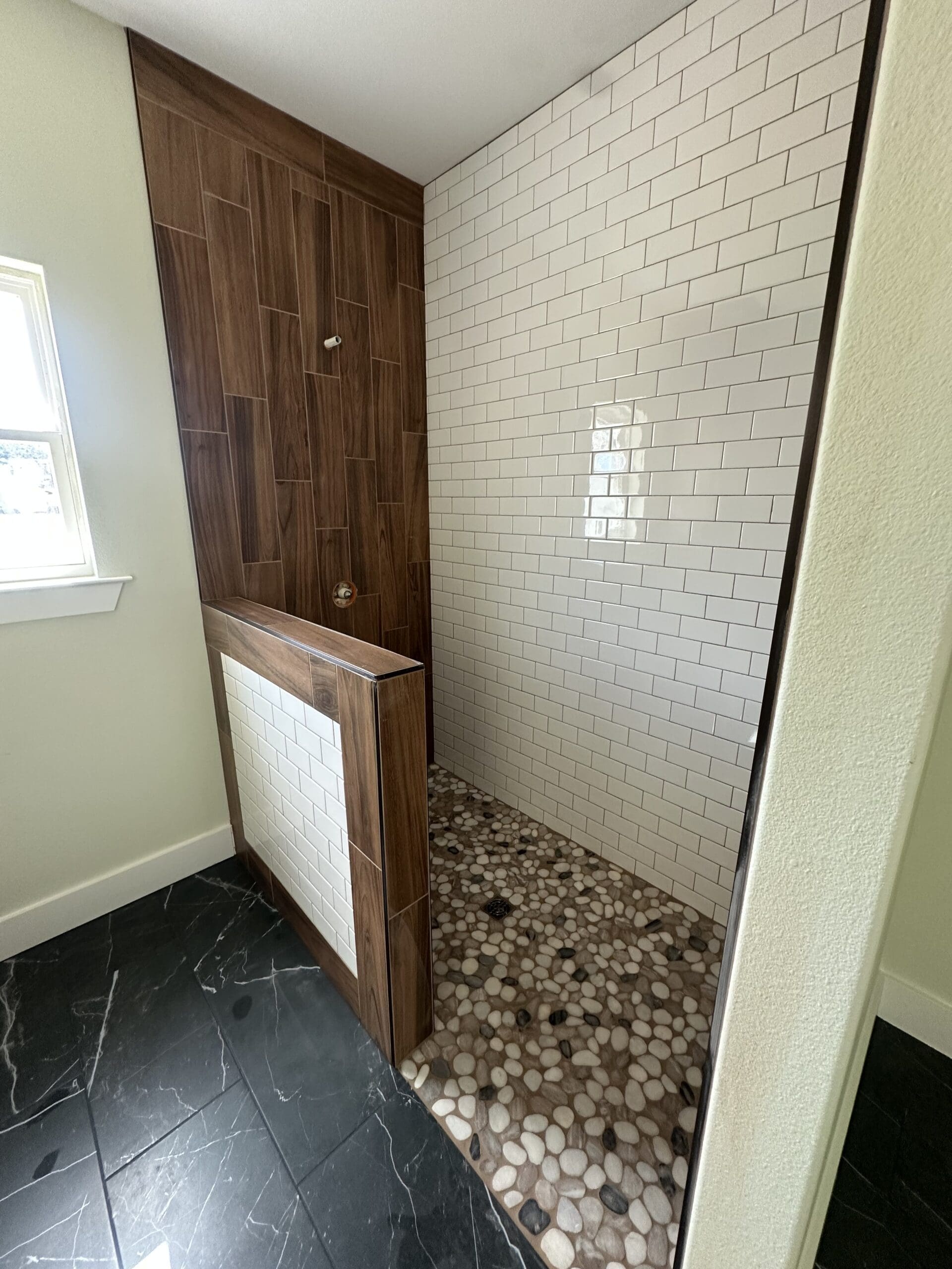 Walk-in shower with white subway tile wall, dark wood-patterned tiles, pebble stone floor, and a short partition wall in a bathroom with black marble flooring—perfect for a custom home or Barndo design.
