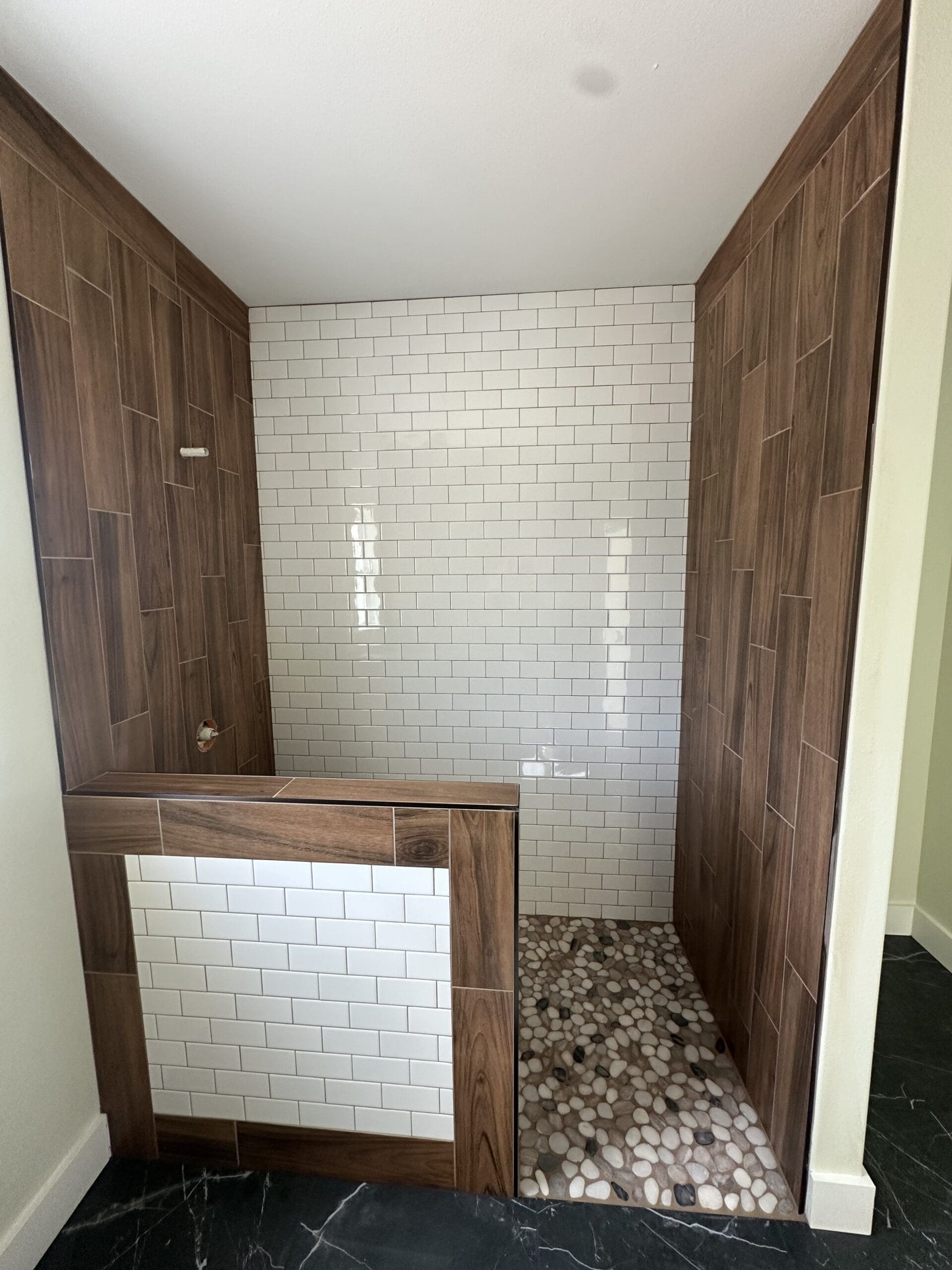 A walk-in shower with white subway tile walls, a half-wall with wood-look tile accents, and a pebble stone floor—perfect for your custom home designed by an expert home builder.