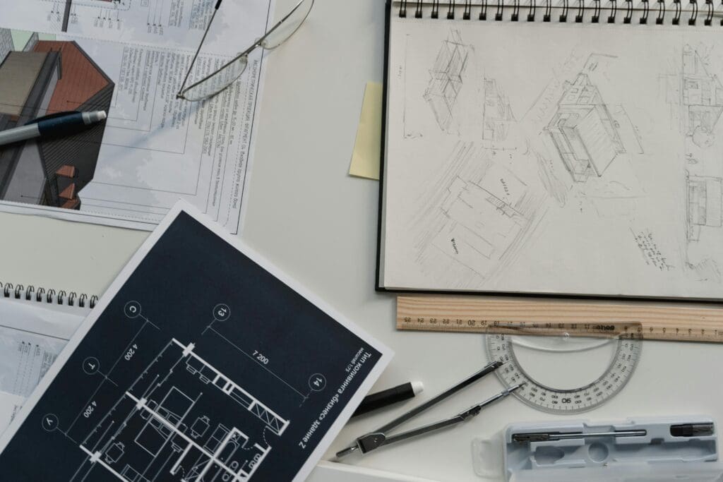 Architectural workspace with blueprints, a sketchbook of building drawings, ruler, protractor, pen, and glasses on a white desk—perfect inspiration for any custom home or home builder project.