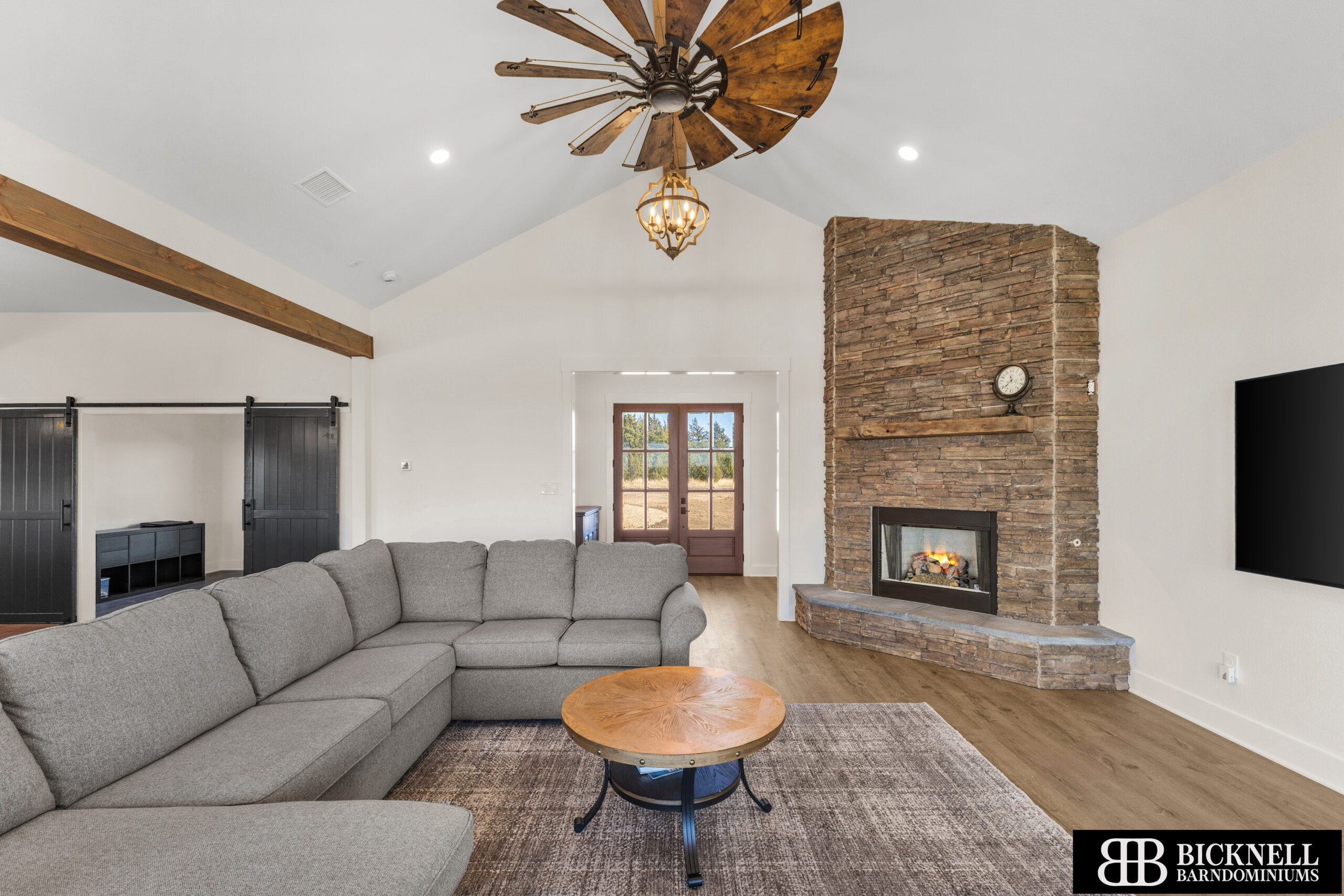 Cozy living room with a large gray sectional sofa, round wooden coffee table, and a stone fireplace with a rustic mantle.