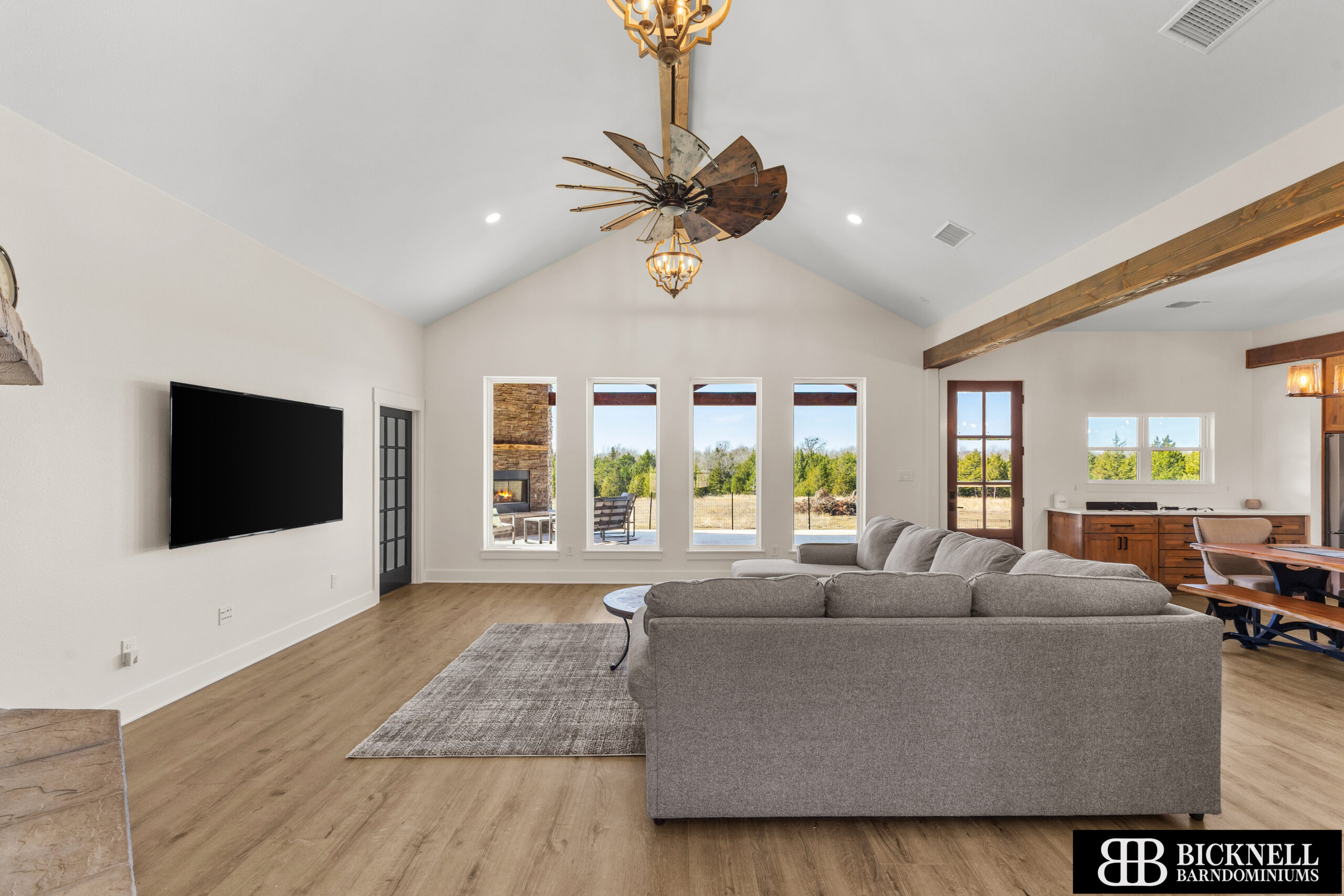 Modern living room with gray sectional sofa, wide windows, and a vaulted ceiling featuring rustic wooden beams.