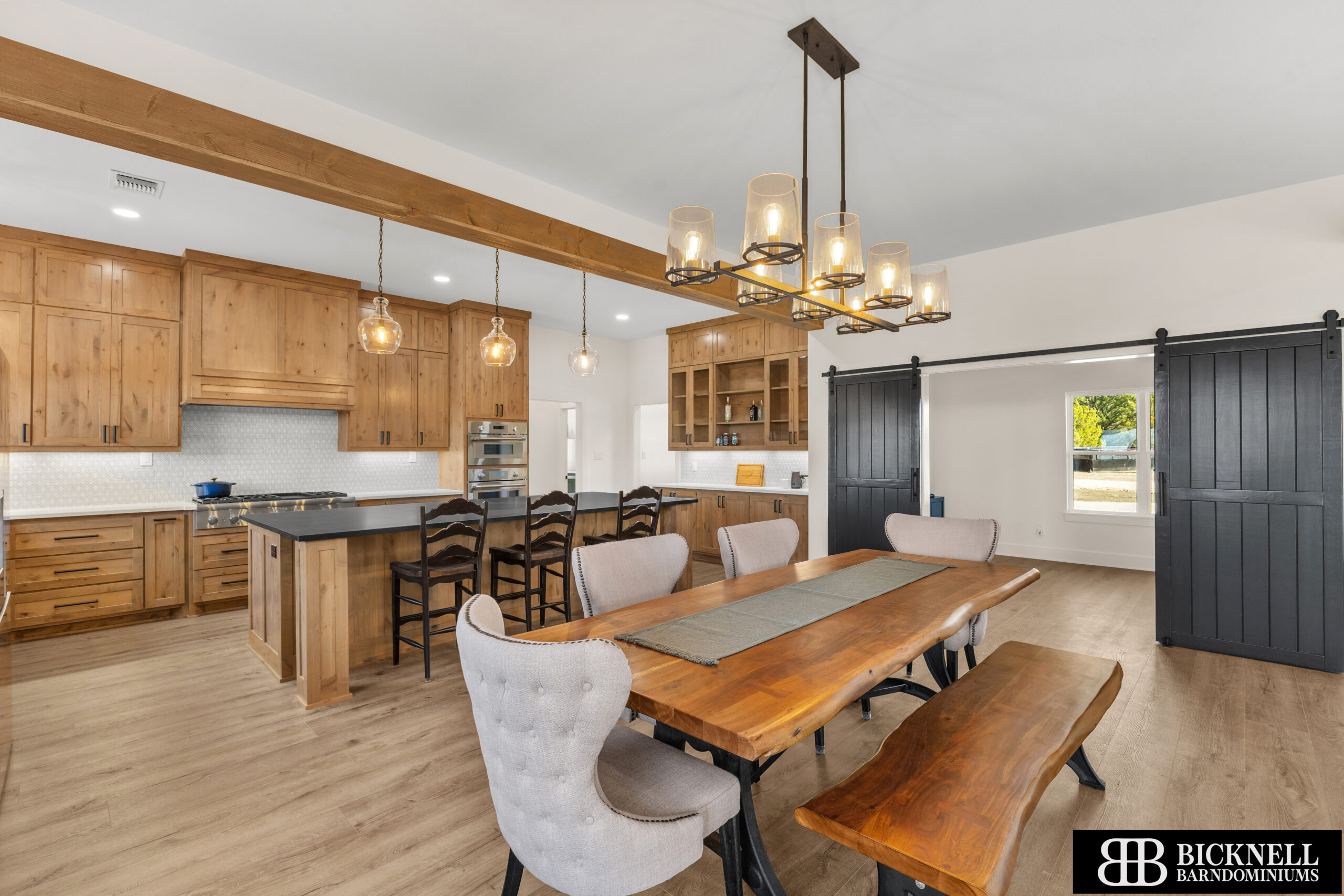 Rustic kitchen with natural wood cabinets, an island, and a dining table under a row of glass pendant lights in a bright open space.