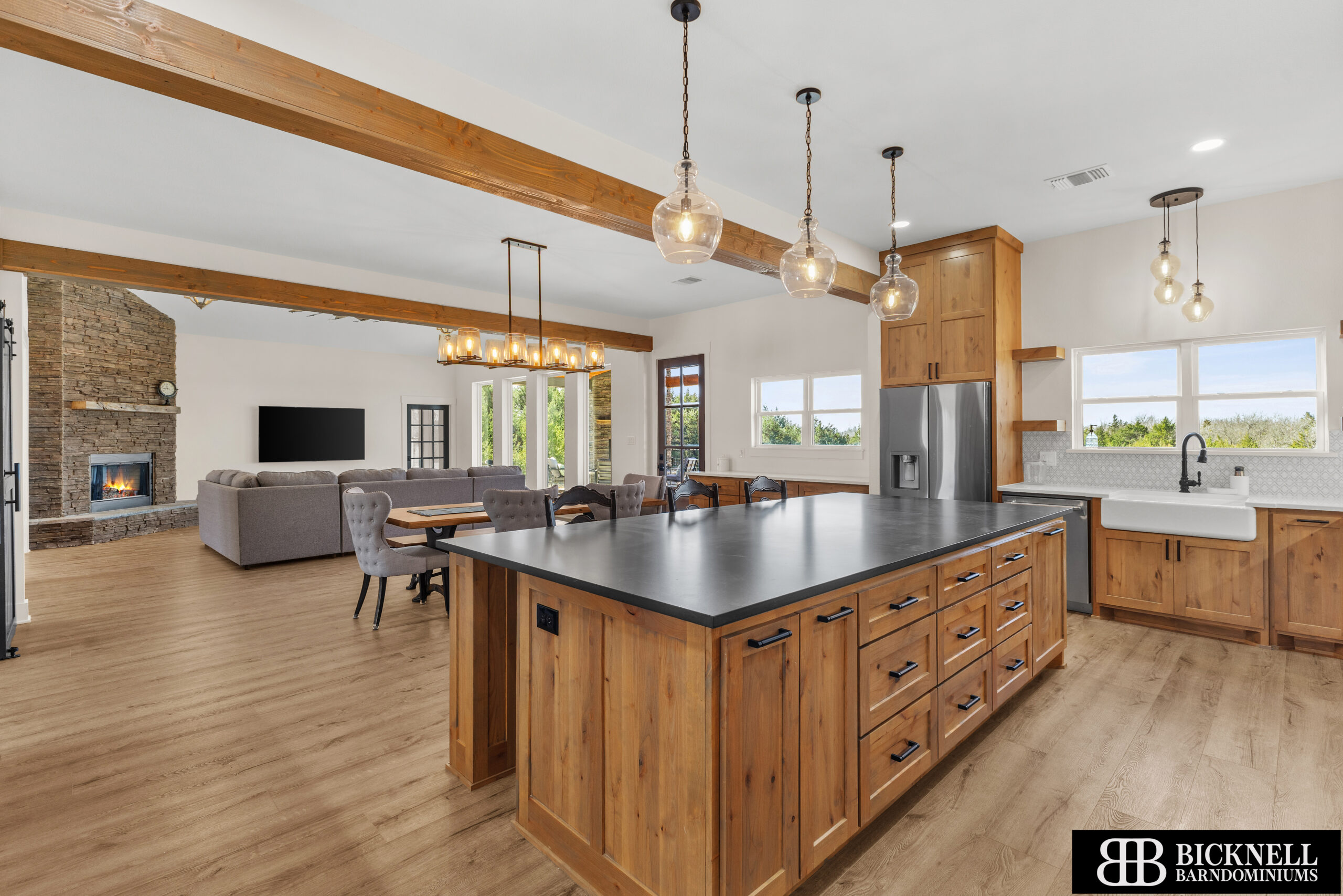Spacious open-concept kitchen and living area with wood flooring, large island, farmhouse sink, modern appliances, and stone fireplace in the background.