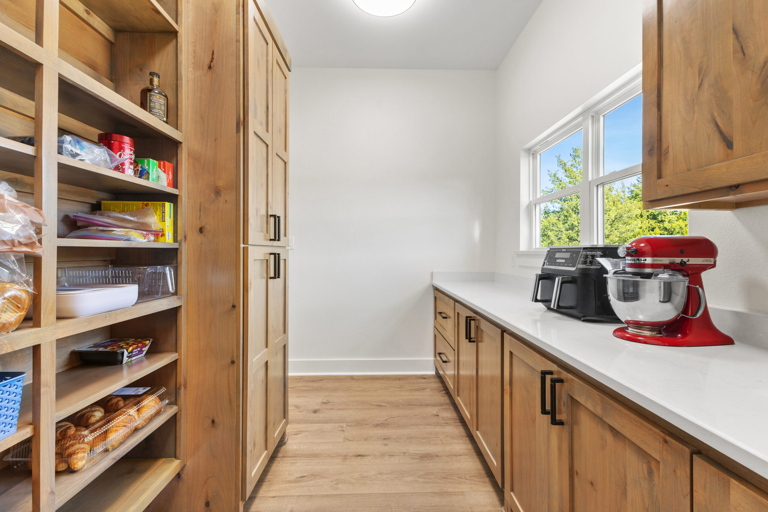 A pantry with wooden shelves and cabinets, countertop appliances including a red mixer and air fryer, and a window letting in natural light.