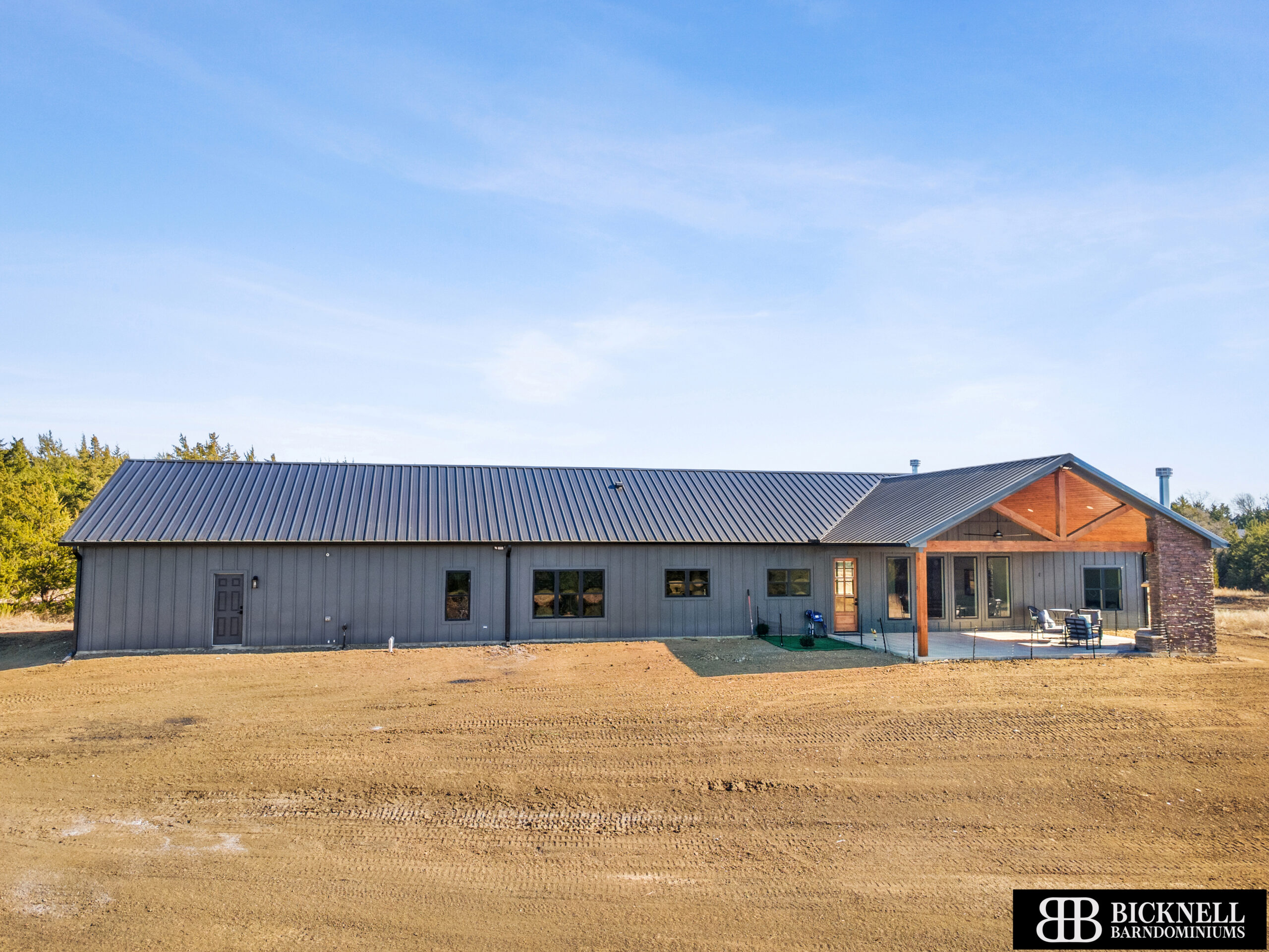 Grey metal-roofed, single-story building under a clear blue sky, with a wooden porch and chairs on the right side.